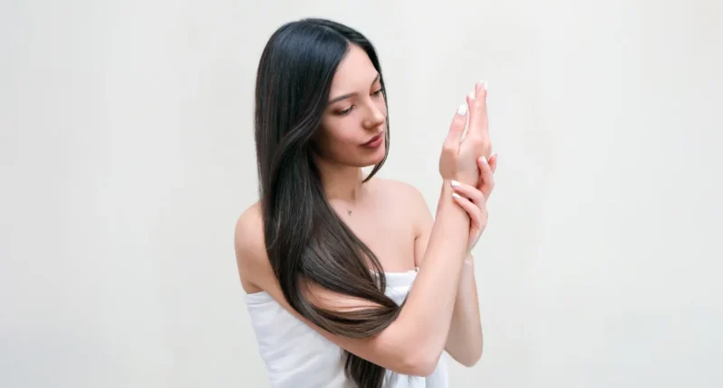 A woman with long, dark hair and flawless skin is gently applying cream to her hands, emphasizing a summer skincare routine for hydration and protection.