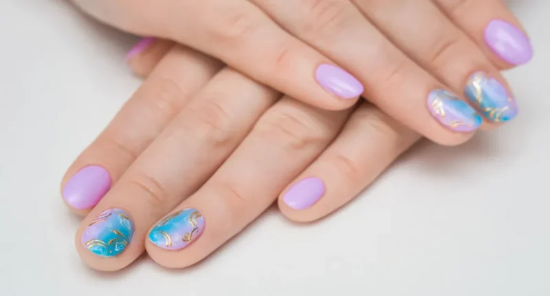 Nails painted in pastel purple with intricate blue and gold designs on two accent nails, perfect for a delicate and artistic summer nail style