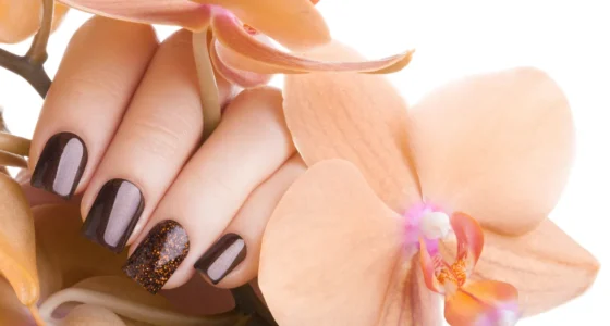 A close-up of a hand with dark brown and glittery accent nails, beautifully displayed against a backdrop of soft peach flowers, highlighting a trendy fall nail design idea.